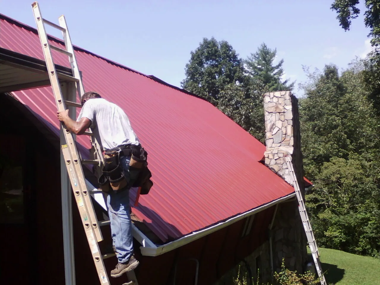 Roofer on a ladder beside a red metal roof and stone chimney — Soffit & Fascia Repair by Roofing Brothers in Penns Grove